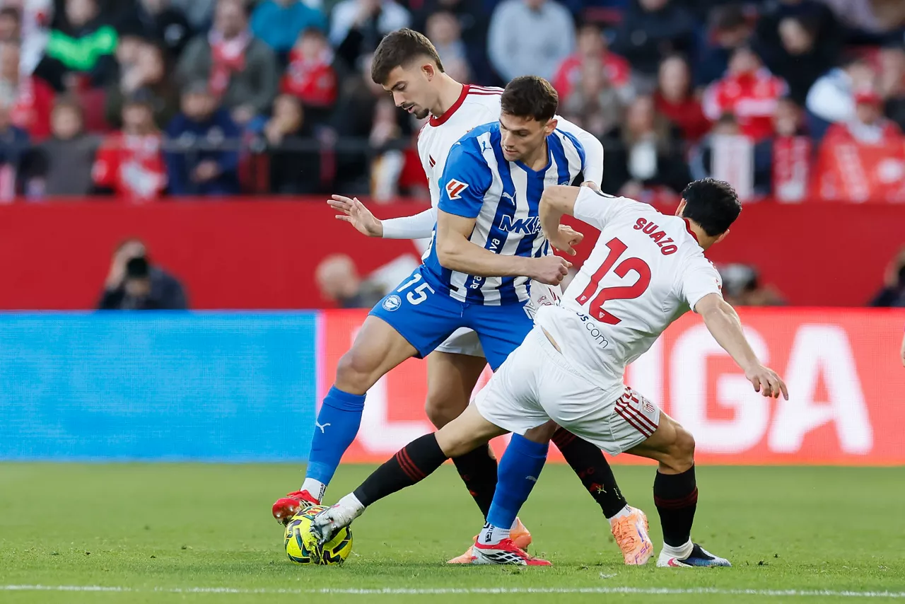 SEVILLA, 14/02/2026.- El delantero argentino del Alavés Lucas Boyé (i) lucha con Kike Salas (i) y con el chileno Gabriel Suazo, ambos del Sevilla, durante el partido de la jornada 24 de LaLiga que Sevilla FC y Deportivo Alavés disputan hoy sábado en el estadio Ramón Sánchez-Pizjuán. EFE/José Manuel Vidal
