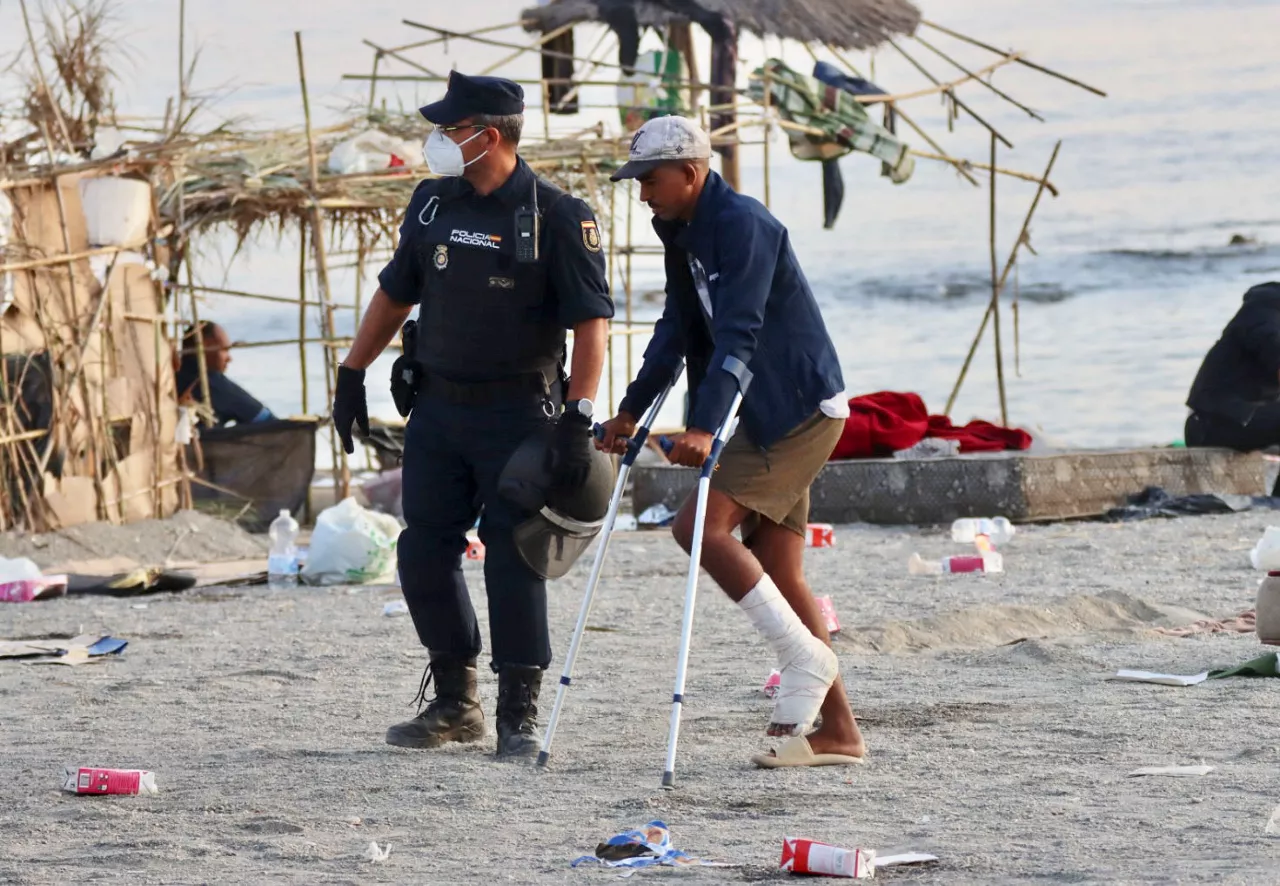 CEUTA, 20/08/2026.- Un policía acompaña a uno de los migrantes durante el desalojo de la playa del Trampolín en Ceuta este jueves. El Gobierno ha habilitado temporalmente varios espacios para la acogida de 1.800 personas migrantes que se encuentran en situación de calle en la playa del Trampolín y en la zona de Loma Colmenar, en Ceuta, que prevé ampliar progresivamente. EFE/Reduan
