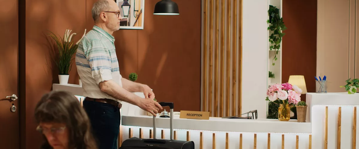 Elderly guest arrives in hotel lobby, greeted by skilled staff members at reception. Senior person carrying luggage for international vacation, preparing for check in, ring service bell.
